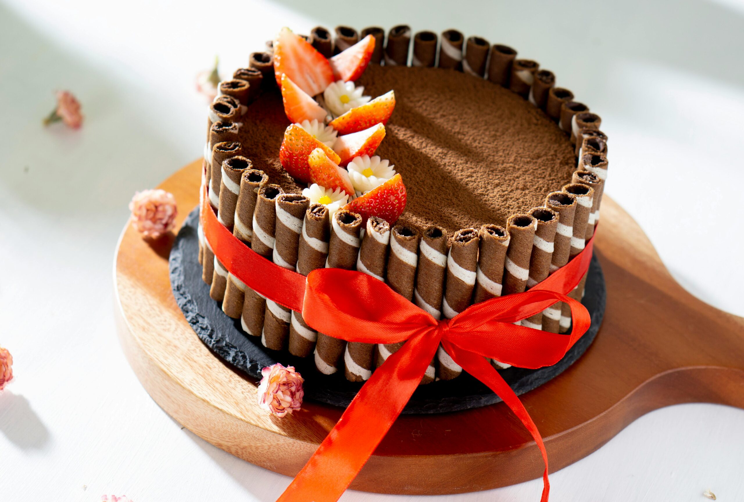 Eggless Moist Chocolate Cake Recipe.Full eggless chocolate cake decorated with wafer rolls, red ribbon, and fresh strawberry slices on a wooden serving board.