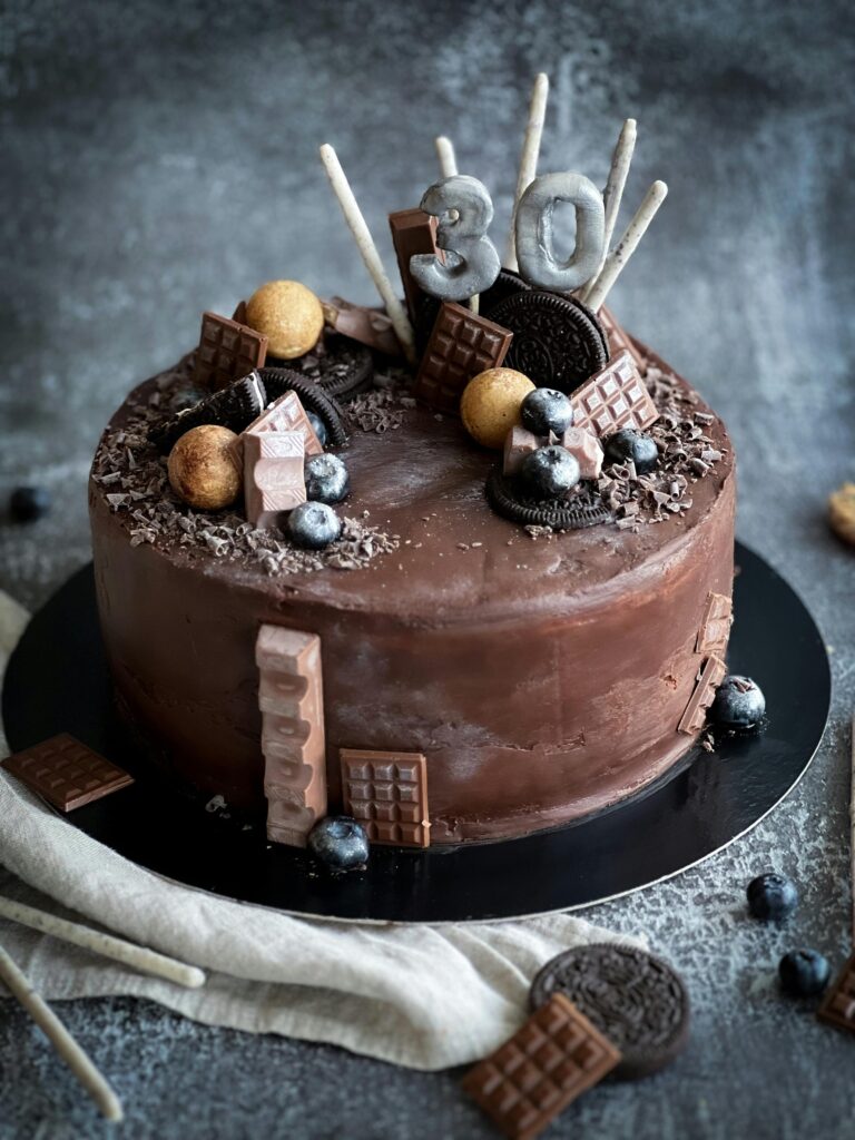 Eggless Moist Chocolate Cake Recipe.Rich dark chocolate cake decorated with mini chocolate bars, blueberries, Oreo cookies, and silver '30' number toppers on a black cake board.