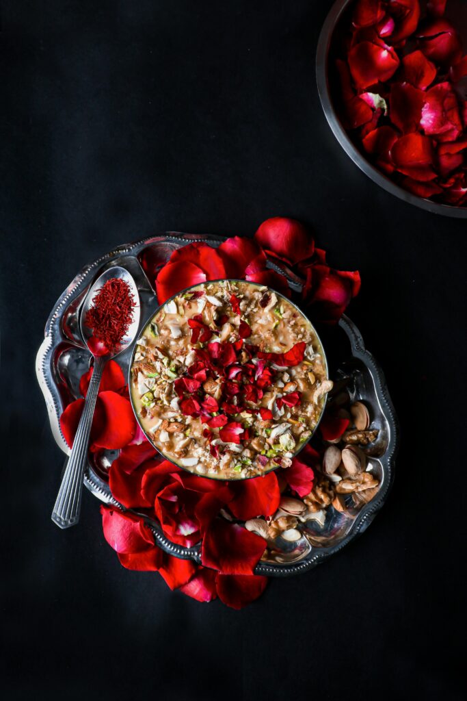 Top view of traditional creamy sheer khurma bowl decorated with fresh red rose petals, chopped pistachios, almonds, and a silver spoon filled with saffron strands