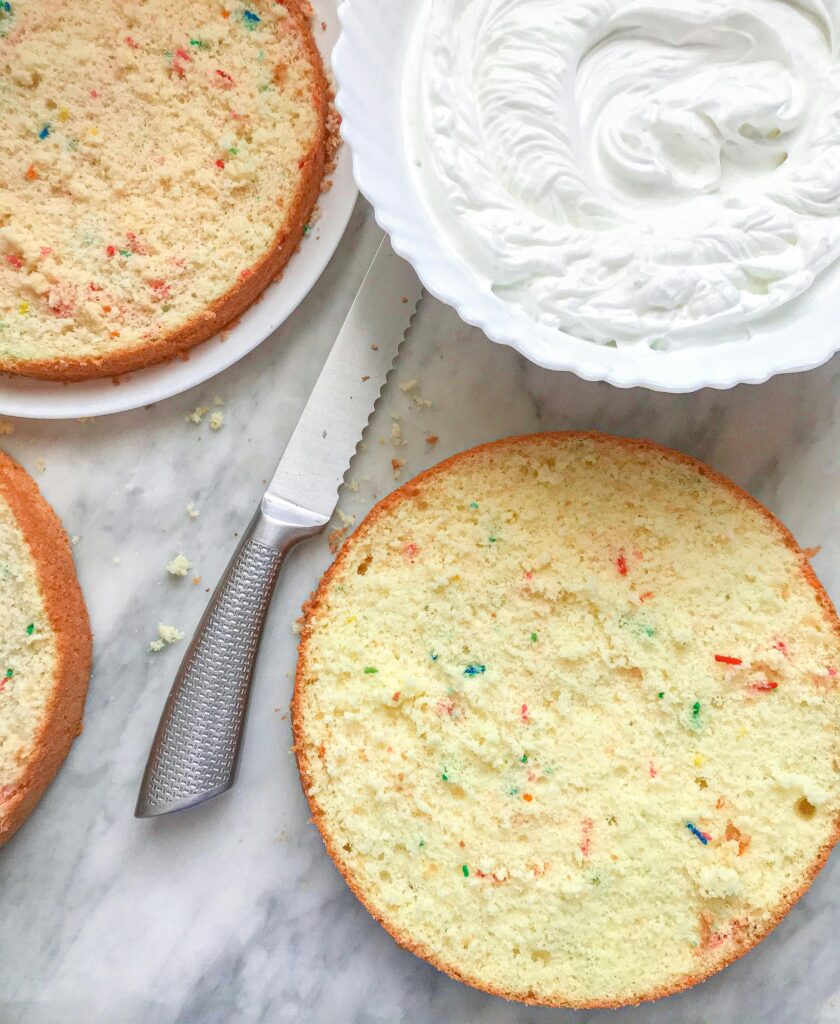 Top view of sliced vanilla sponge cake layers with colorful sprinkles and a bowl of fresh whipped cream frosting on a marble surface.