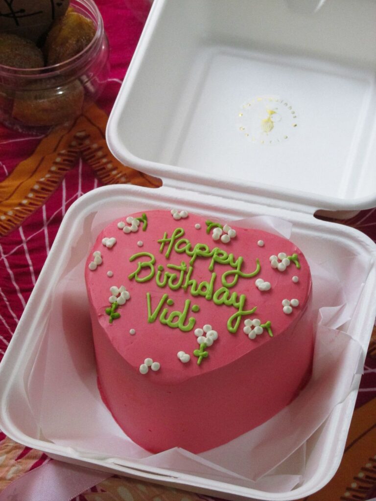 A pink heart shaped minimalist bento cake for beginners with happy birthday green lettering inside a white lunchbox.